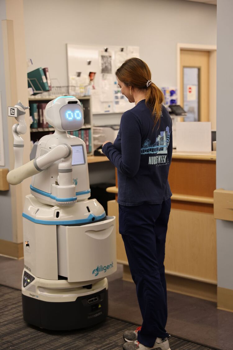 ‘Moxi’, the Diligent Robot (legless) in a hospital setting stands in front of an employee with the open drawer in the center of the robot.