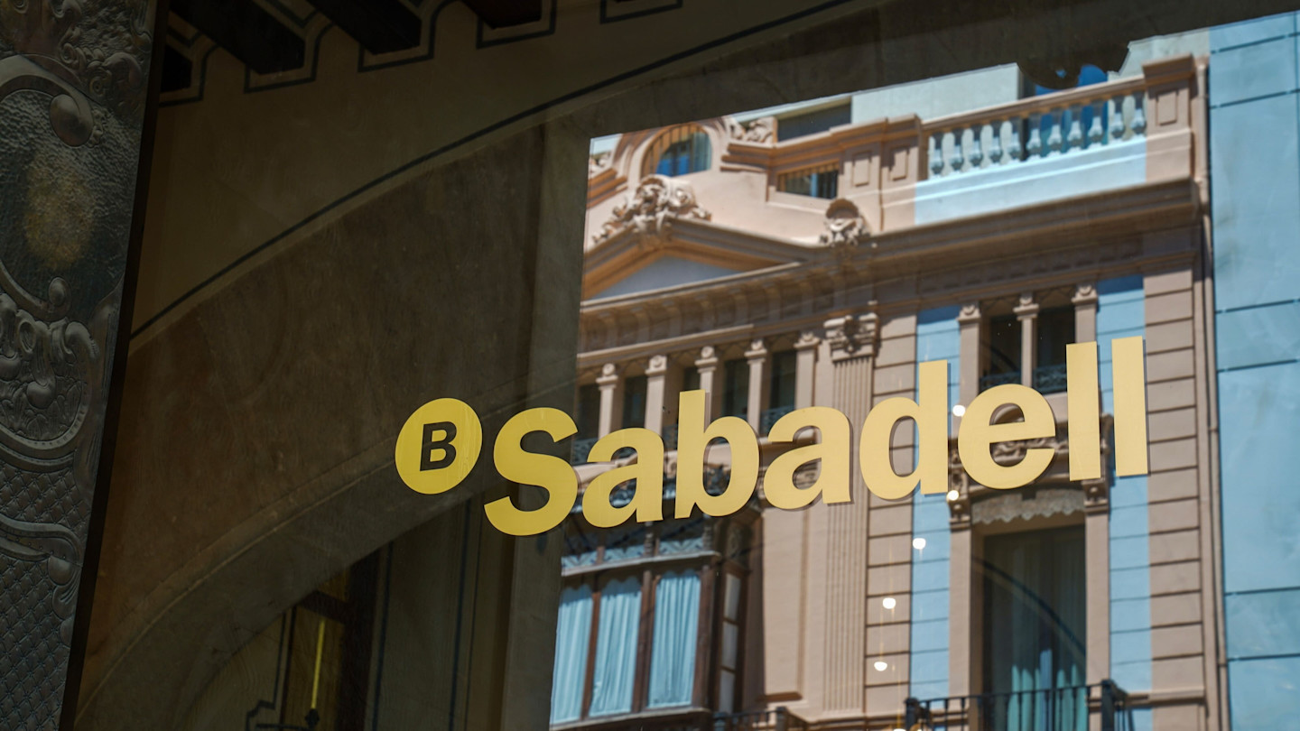 Banco Sabadell logo on a glass window, with the reflection of a historic building facade in Valencia.