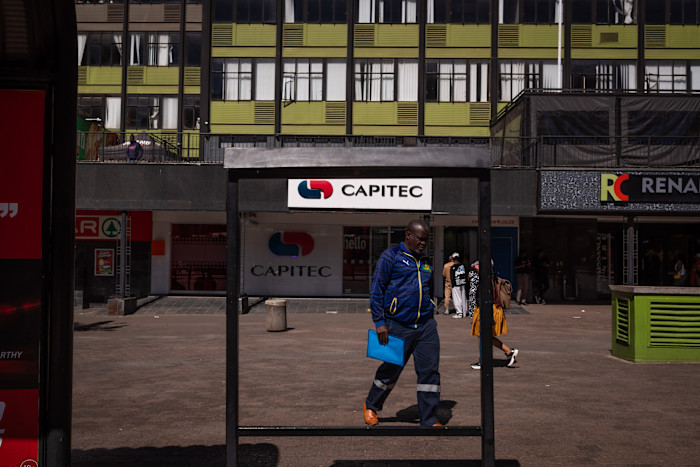A man in a blue jacket walks past a Capitec bank sign in Ghandi Square, with several people and shops in the background.