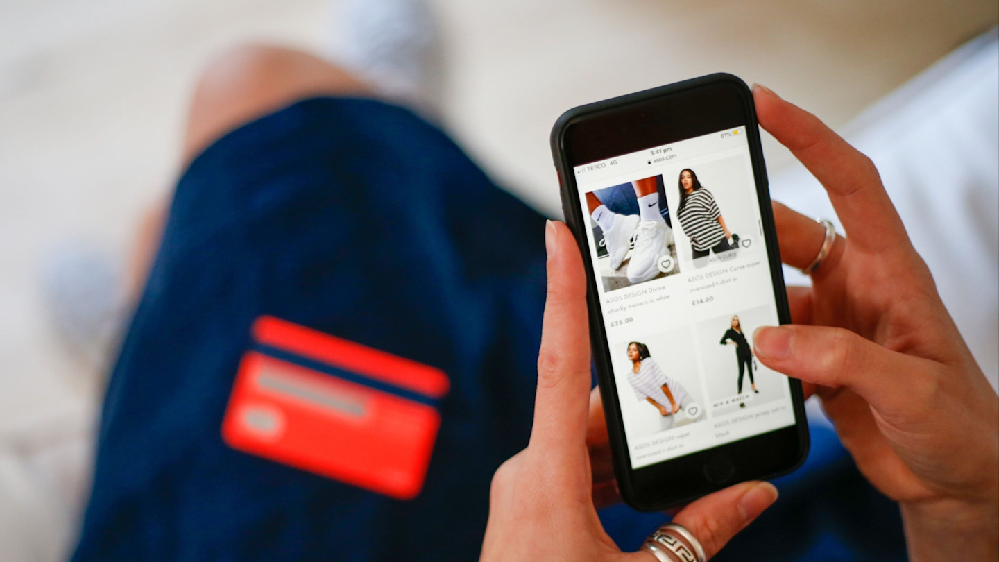 A woman holds a phone and browses clothing items on the Asos website, with a red bank card in her lap.