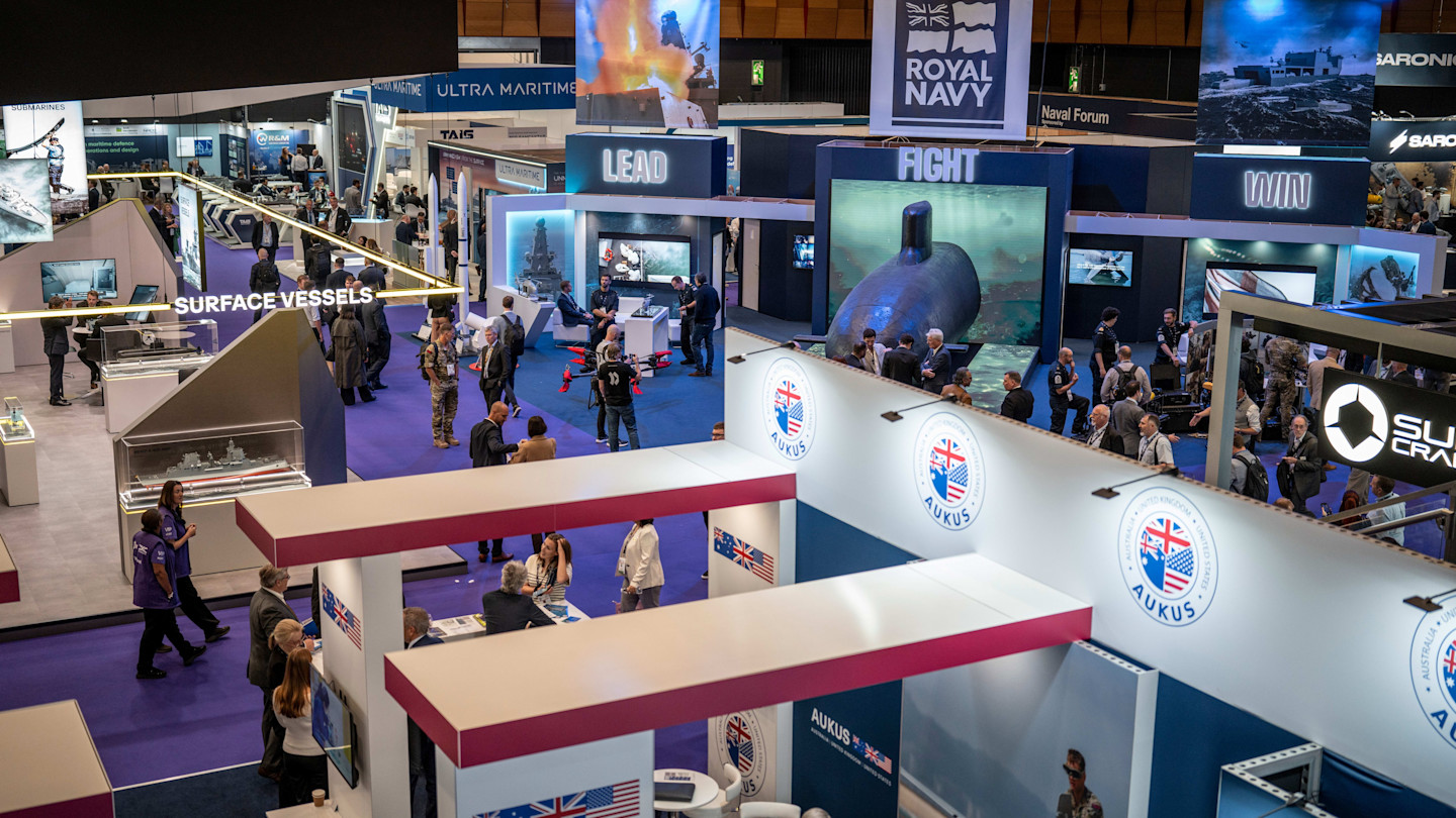 Crowds of visitors walk among exhibitions and military displays, including models of ships and submarines, at the DSEI event.