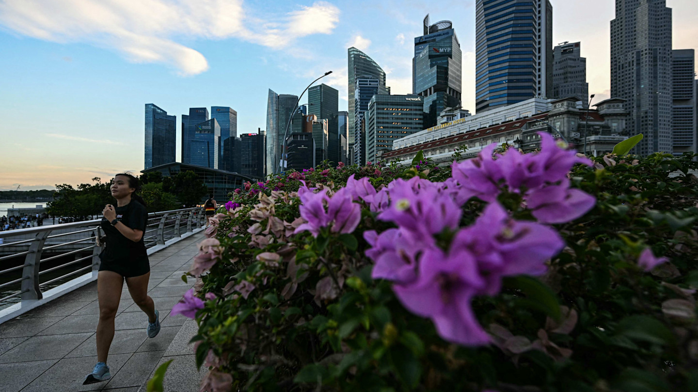 A woman jogs on a riverside path with purple flowers in the foreground and the Singapore city skyline in the background.