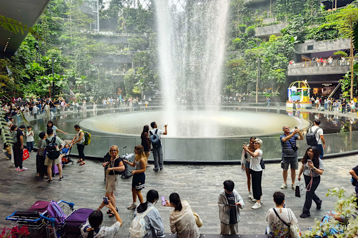 Crowds of people taking photos and admiring the HSBC Rain Vortex, the world's tallest indoor waterfall, surrounded by lush greenery inside Jewel Changi Airport.