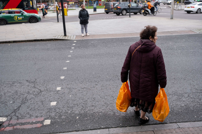 Woman crossing a street carrying two orange Sainsbury's bags, with traffic and pedestrians in the background at Elephant and Castle.