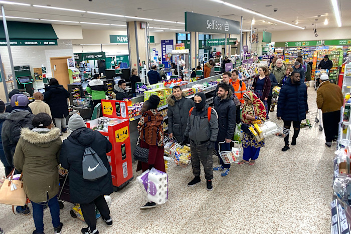 Shoppers stand in a long queue at the checkout area of a supermarket, many holding baskets and toilet paper packs.
