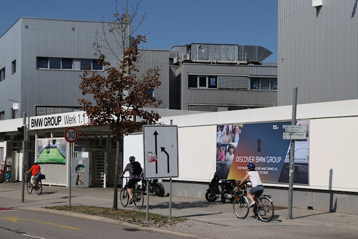 People ride bicycles past the entrance to a BMW factory, with a sign reading ‘BMW GROUP Werk 1.1’ visible on the building.