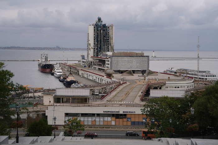 Odesa ship terminal with a tall hotel building in the centre showing severe damage and broken windows from a rocket attack. Several ships are docked nearby