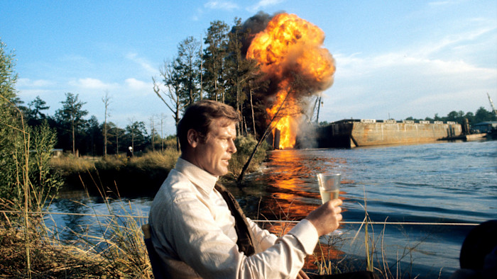 Roger Moore sits by a dam on a river or lake in a cream shirt holding a drink, with a large explosion and fire in the background on the set of the film ‘Live and Let Die’.
