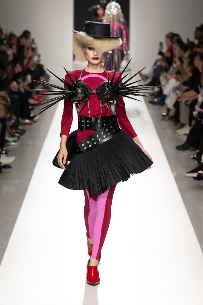 A model walks the runway in a structured magenta and black outfit with dramatic spiked accents on the chest, a wide black belted skirt, and a black hat at Junya Watanabe’s Spring Summer 2027 show.