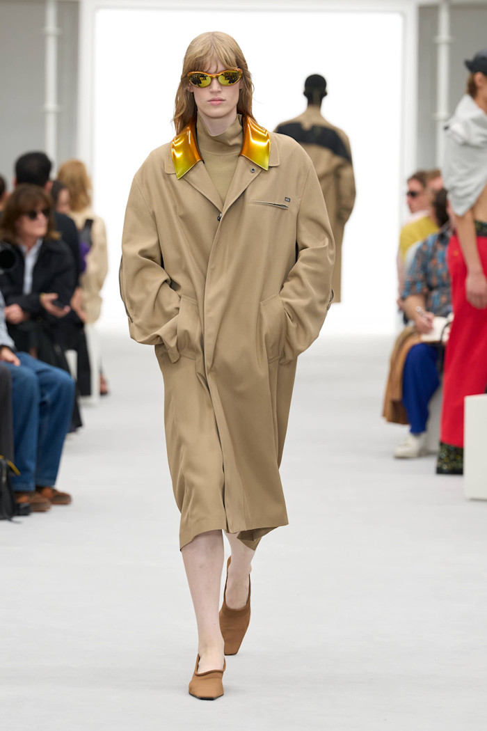 A model in orange-tinted sunglasses walks the runway in an oversized beige coat with a metallic gold collar and brown shoes.