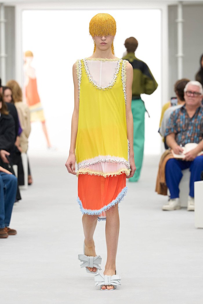 A model walks the runway in a layered yellow, pale pink and orange dress with fringed trim, a yellow beaded headpiece and bow-front sandals.