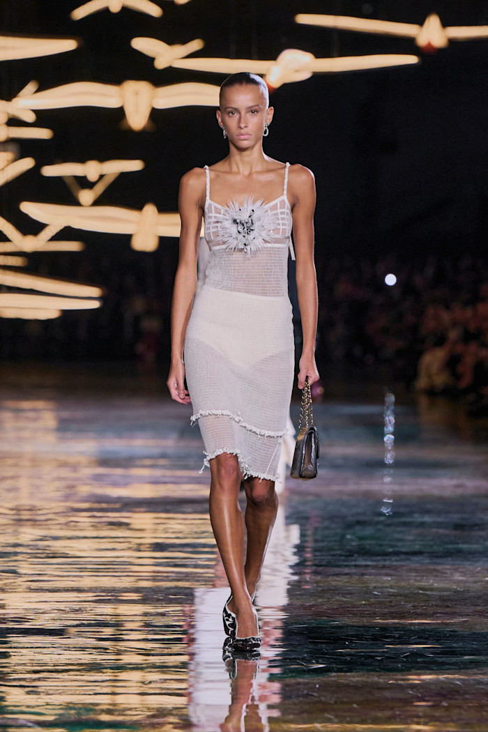 A model walks the runway in a sheer white cardigan and fitted knee-length skirt, with a feathered floral detail across the chest.