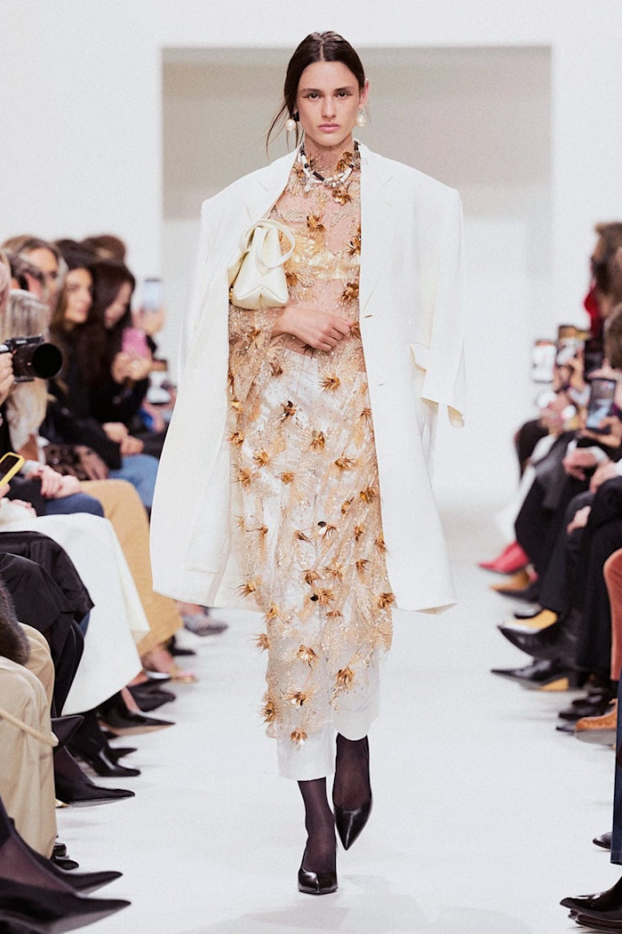 A model walks the runway in an oversized white blazer over a sheer gold embellished dress, carrying a small cream handbag.