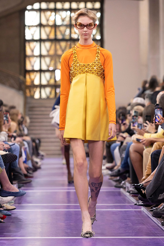 A model in sunglasses walks the runway in an orange long-sleeved top beneath a yellow mini dress with a gold embellished bodice.