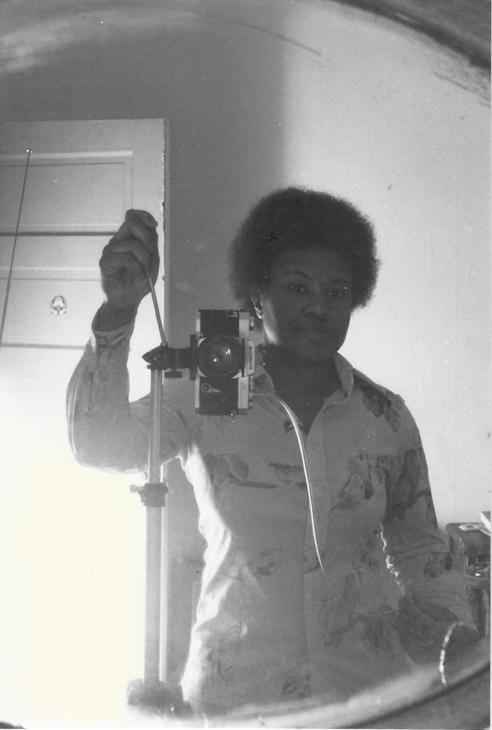 Beverly Buchanan takes a black-and-white self-portrait in a mirror, holding a Konica camera on a tripod