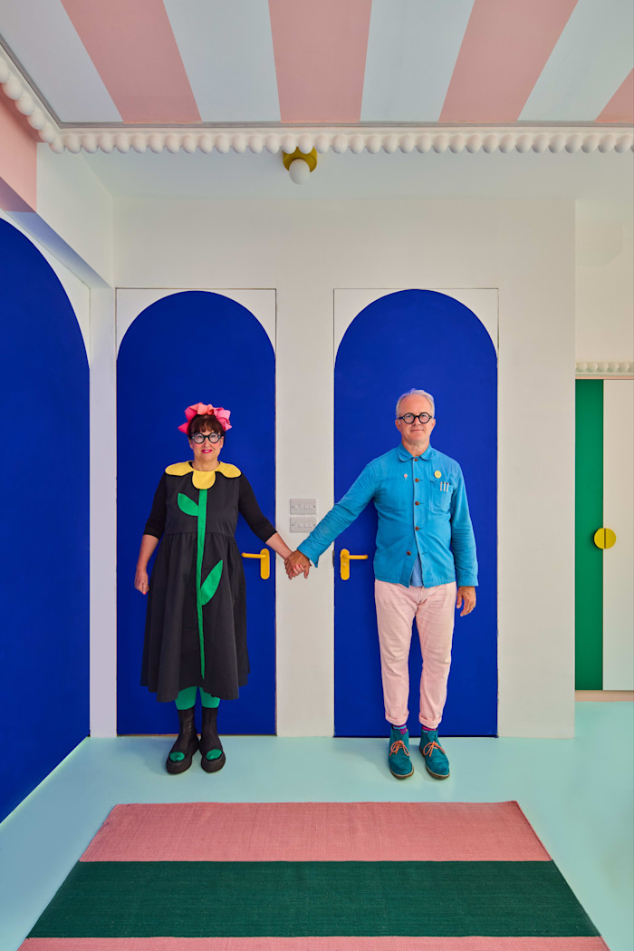 Two people stand holding hands in a brightly coloured room with bold blue arched doors and playful, pastel decor.