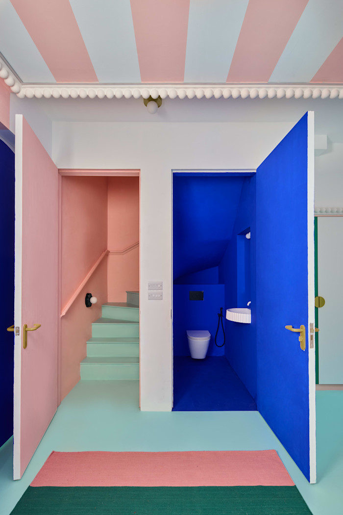 A pastel pink stairwell with mint green steps is next to a vivid blue bathroom with a white toilet and small sink, both with open doors.