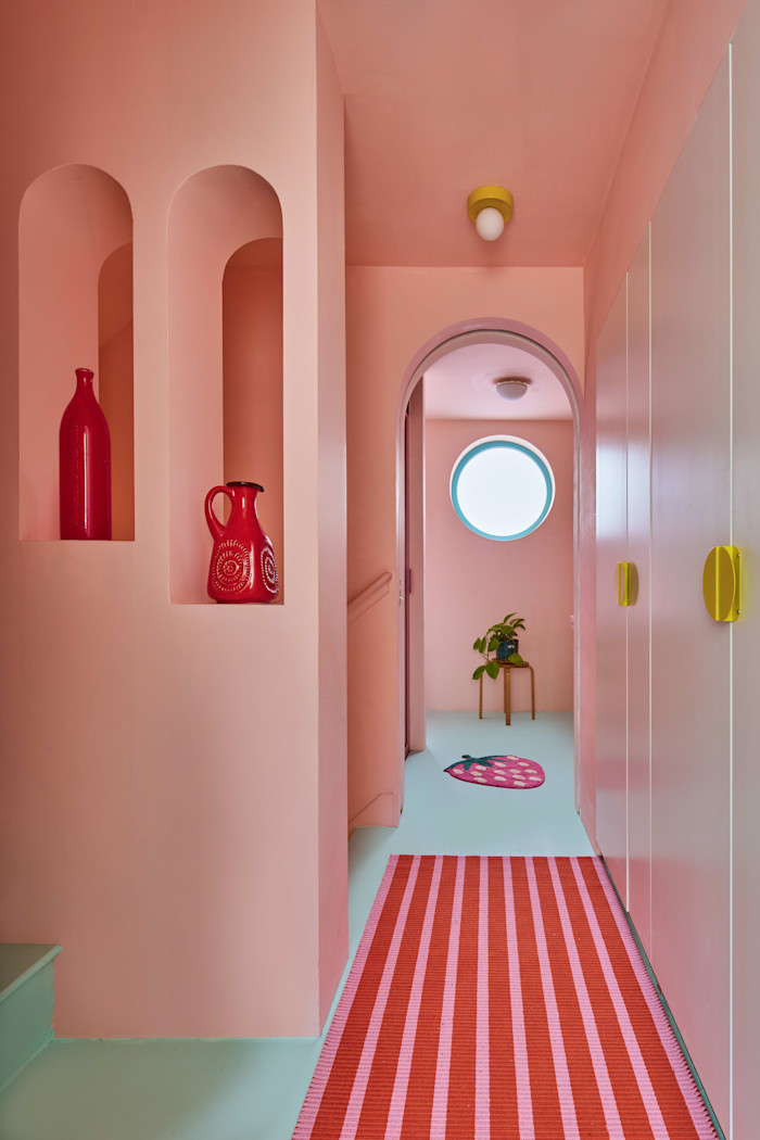 A hallway painted in pastel pink with arched doorways, built-in shelves holding red vases, a striped red and pink rug and a circular window at the end.
