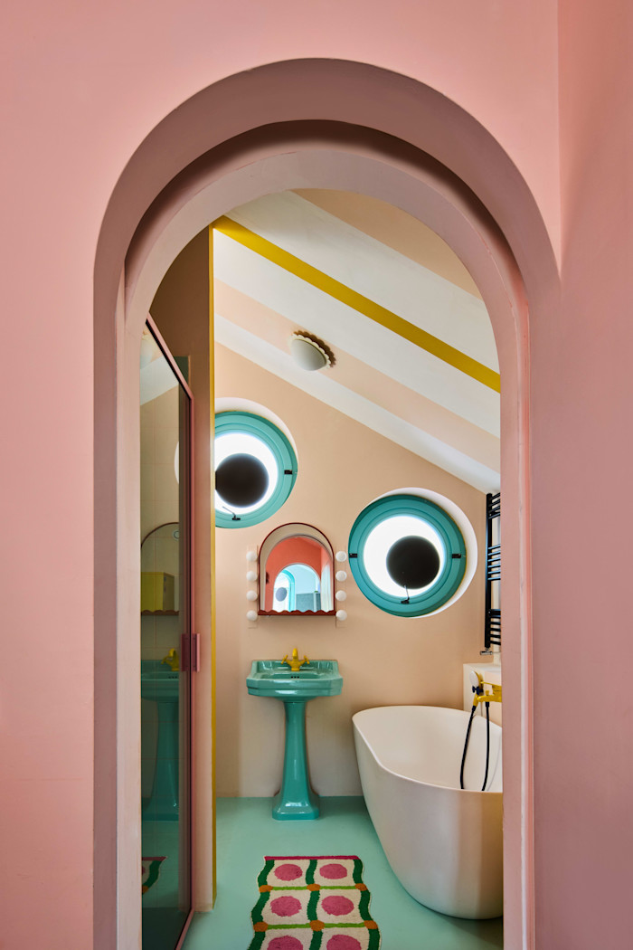 Bathroom with pastel pink walls, turquoise pedestal sink, freestanding white tub, round porthole windows, and colorful geometric rug.