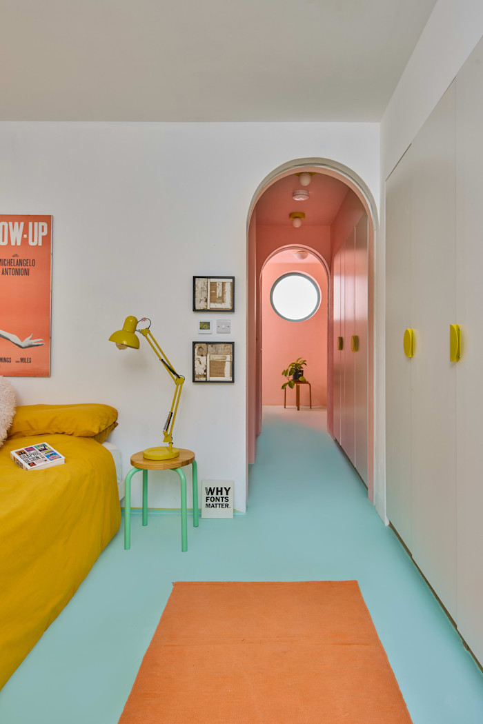 Bedroom with a yellow bedspread, yellow desk lamp, and orange rug, leading to a pink hallway with a circular window and mint-green floor.
