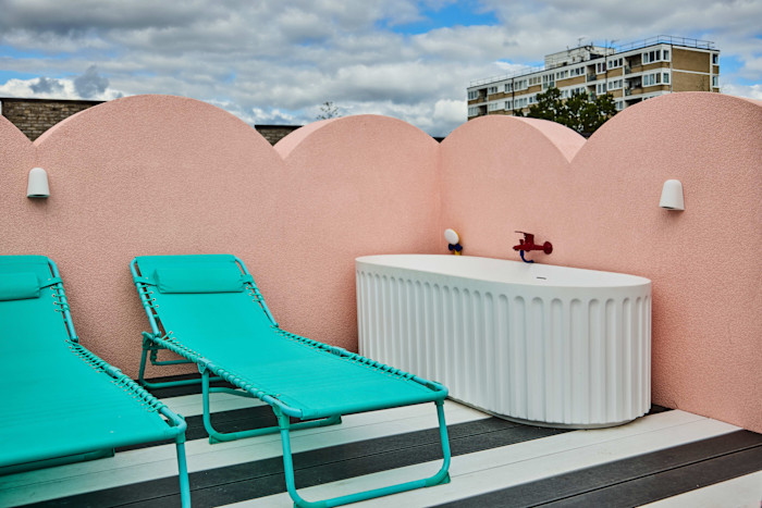 Two turquoise lounge chairs and a white freestanding bathtub sit on a deck against a pink scalloped wall under a cloudy sky.
