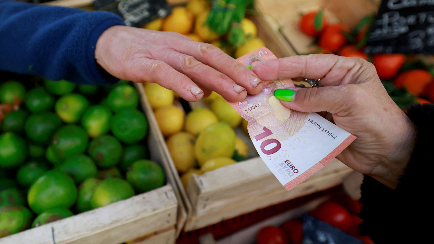 A person hands a €10 banknote to another person at a market stall with lemons and limes in the background.