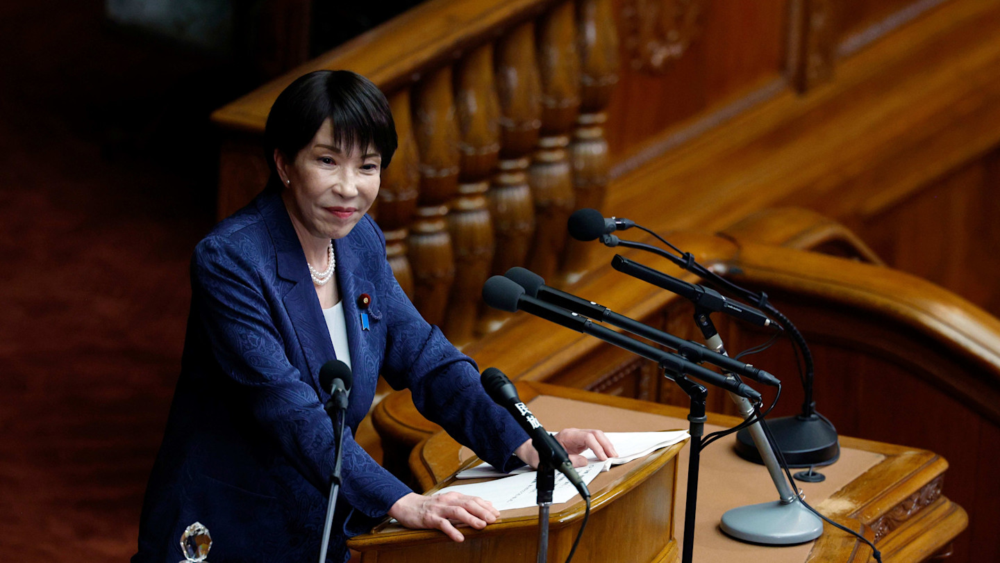 Japanese Prime Minister Sanae Takaichi stands at a podium delivering a speech in parliament, with multiple microphones in front of her.