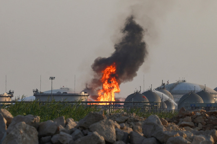Flames and thick black smoke rise from an Aramco oil facility, with industrial storage tanks visible in the background.