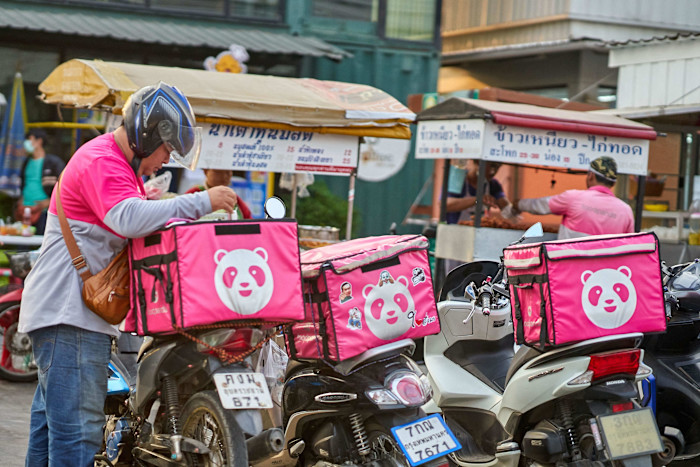 A person in a pink and grey uniform with a helmet prepares a food delivery bag next to motorcycles with large pink Foodpanda boxes.