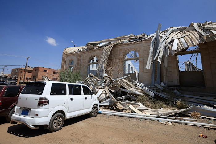 A sports hall with a collapsed metal roof and severe structural damage after an air strike, with parked cars in front.