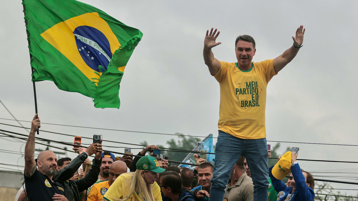 Flavio Bolsonaro stands on a vehicle waving with both hands during a campaign rally, surrounded by supporters and a large Brazilian flag.