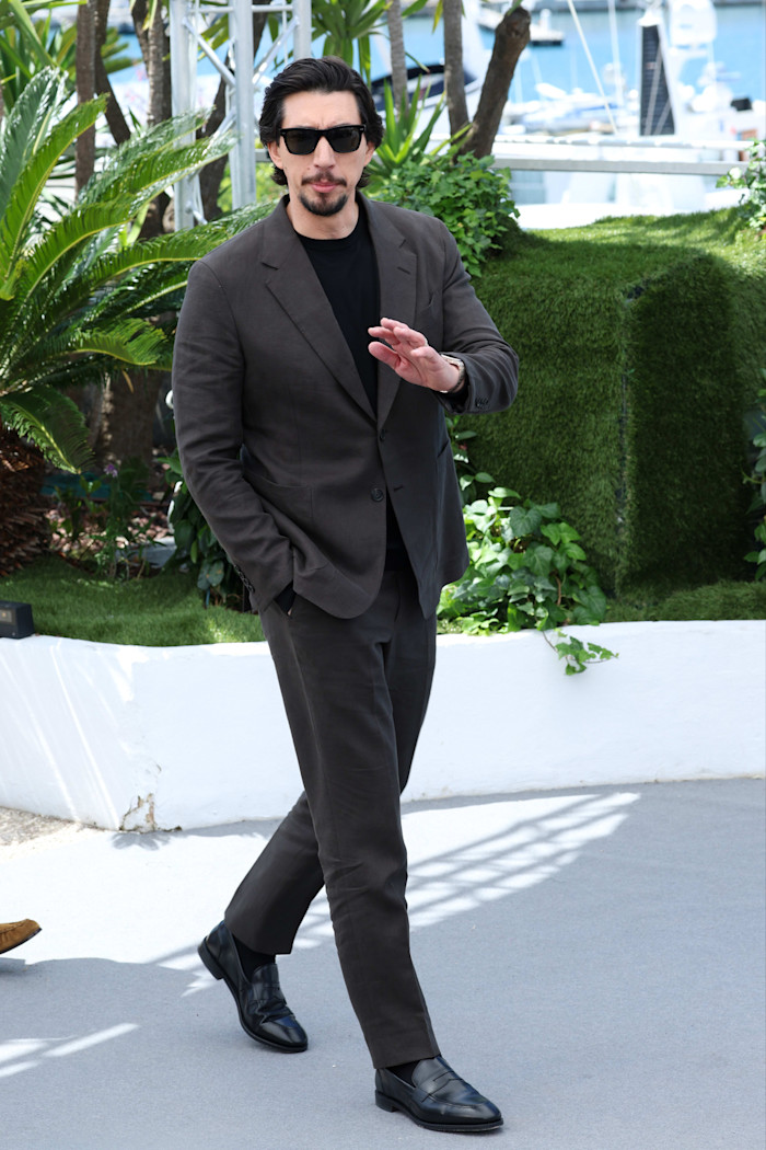Actor Adam Driver walks outdoors on a terrace by a marina in Cannes in a dark suit and sunglasses, raising a hand.