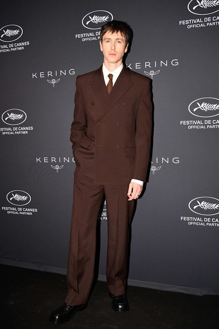 Actor Harris Dickinson poses in a brown double-breasted suit in front of hoardings advertising the Kering brand and Cannes Film Festival.