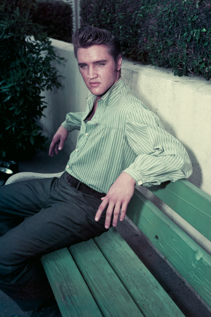 Elvis Presley sits on a green bench, wearing a striped green and white shirt and dark trousers, looking towards the camera.