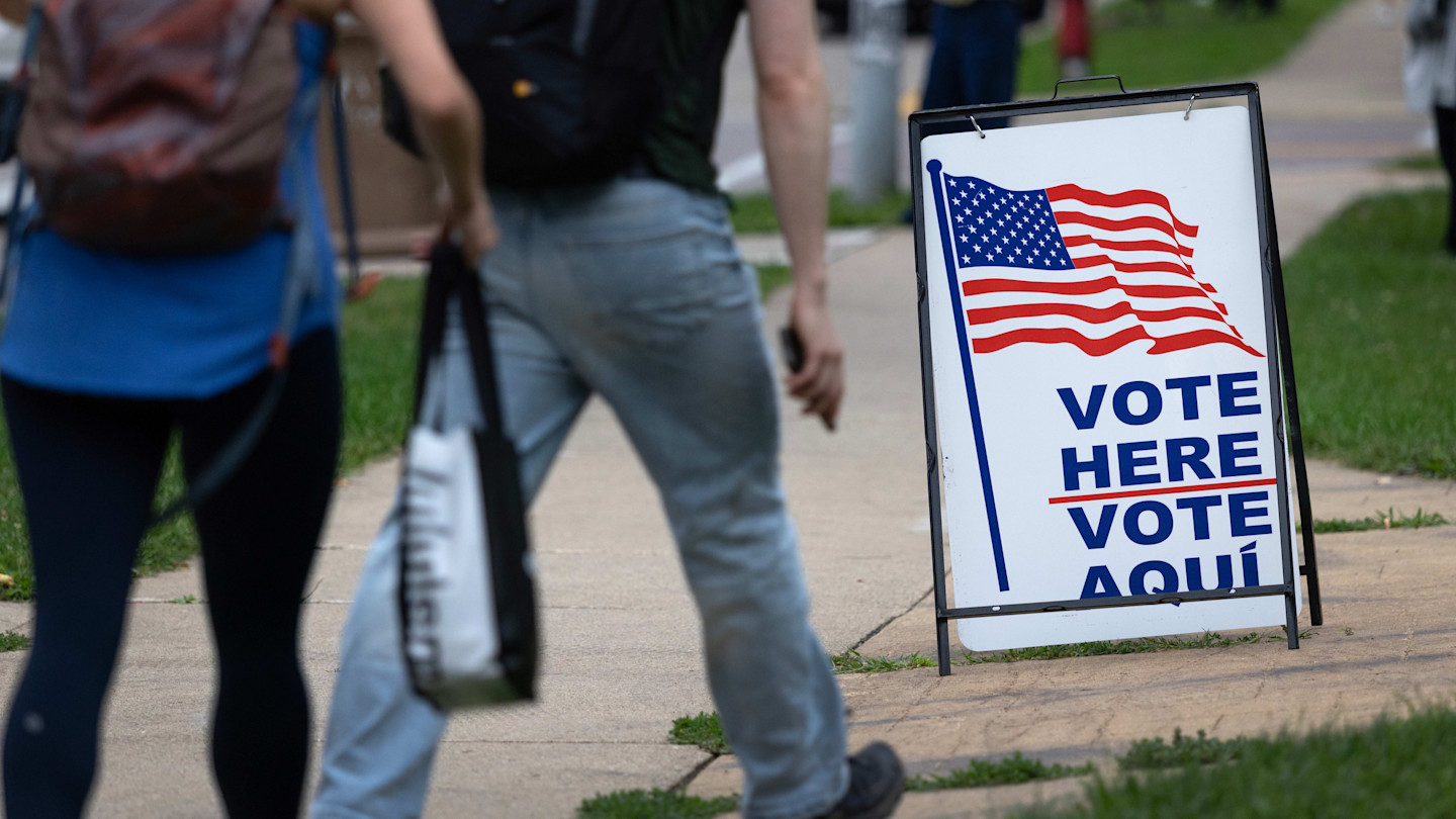 People walk past a sidewalk sign with an American flag that reads “Vote Here Vote Aquí” outside a polling location.
