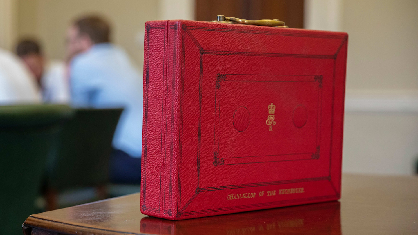 A red briefcase labelled "Chancellor of the Exchequer" sits on a table, with blurred people in the background.