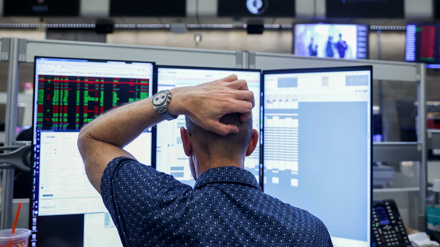 A trader with a hand on his head looks at multiple monitors displaying financial data on a trading floor.