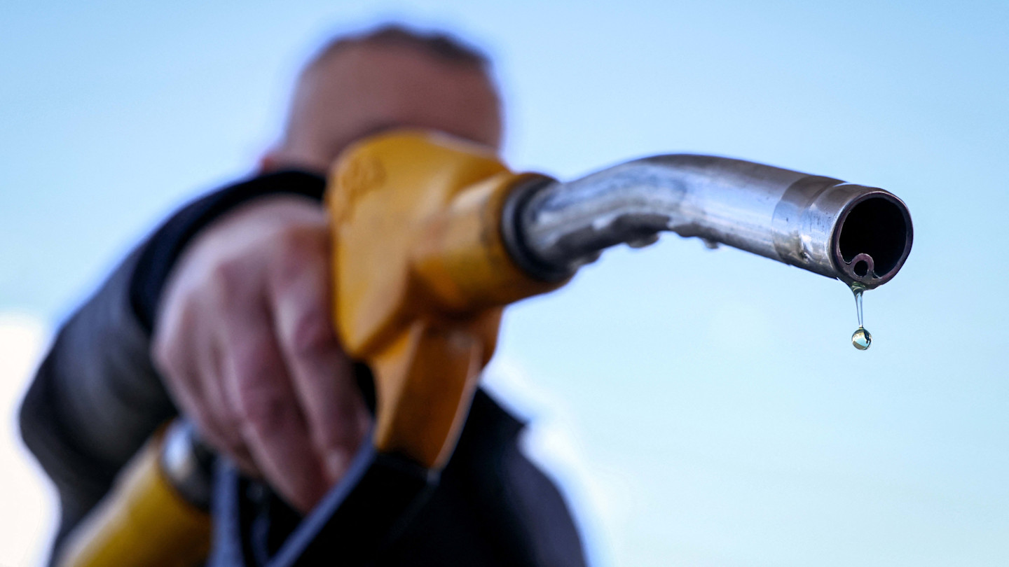 A close-up of a hand holding a yellow petrol pump nozzle with a drop of petrol falling from the tip.