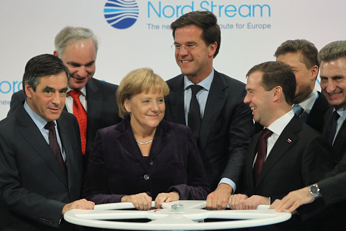 Francois Fillon, Matthias Warnig, Angela Merkel, Mark Rutte, Dmitry Medvedev, and Guenther Oettinger stand together smiling as they hold a large wheel at the Nord Stream pipeline ceremony.