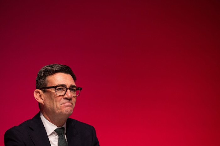 Andy Burnham sits against a plain red background, wearing glasses, a dark suit, white shirt, and tie.