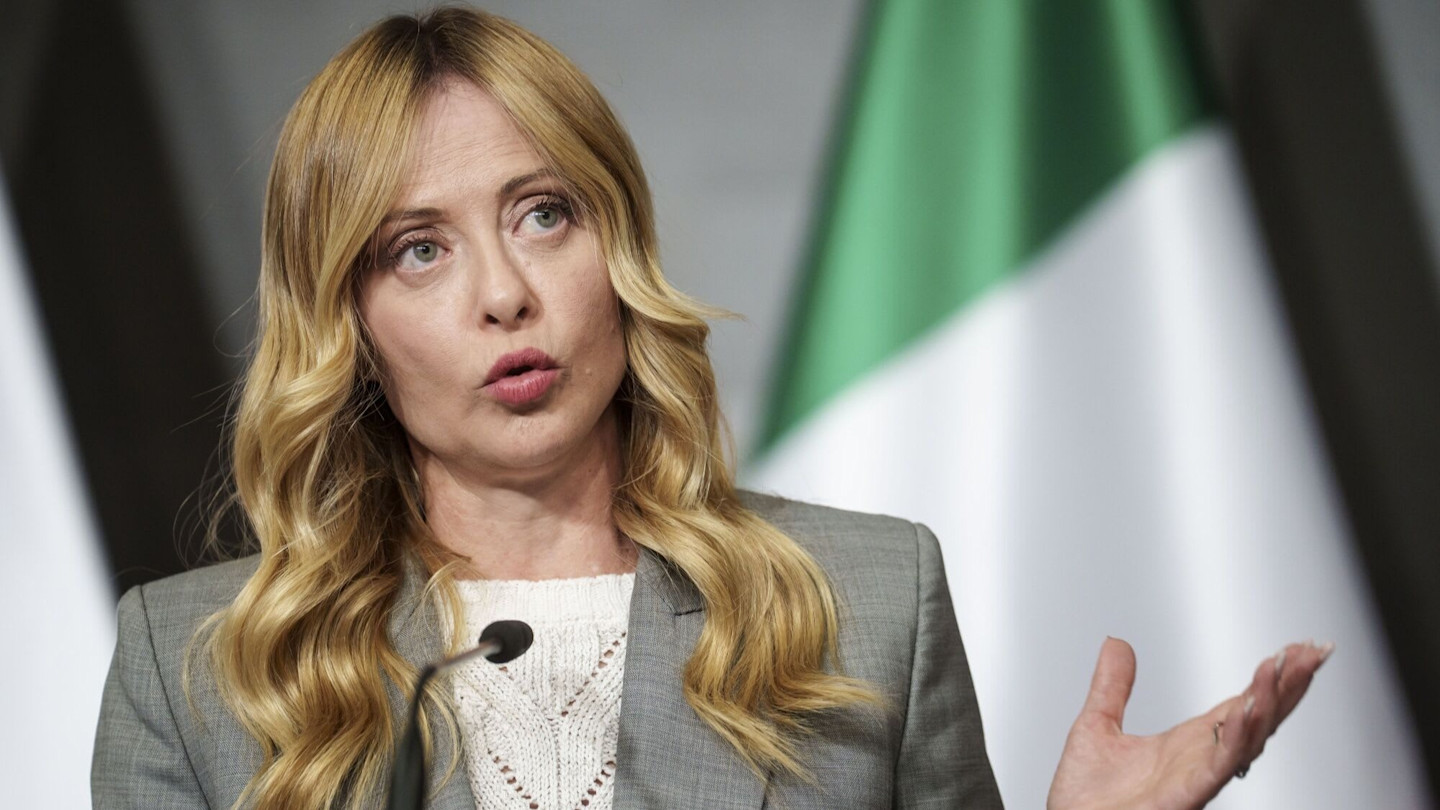 Giorgia Meloni gestures while speaking at a news conference, with a blurred Italian flag in the background.