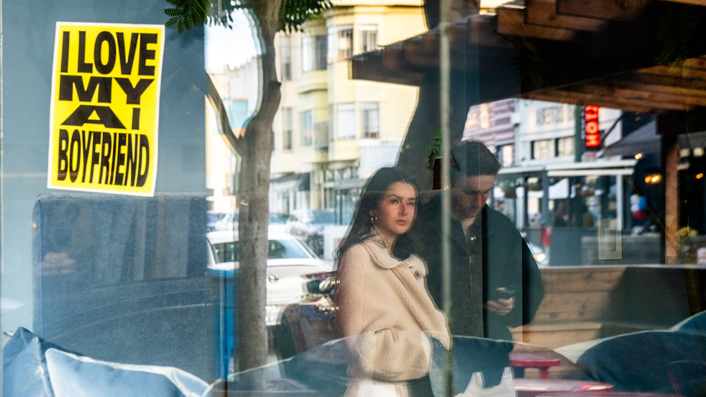 A yellow poster reading “I love my AI boyfriend” is displayed in a window, with a passing couple reflected in the glass — a young woman looking at the window and a young man looking at his phone.