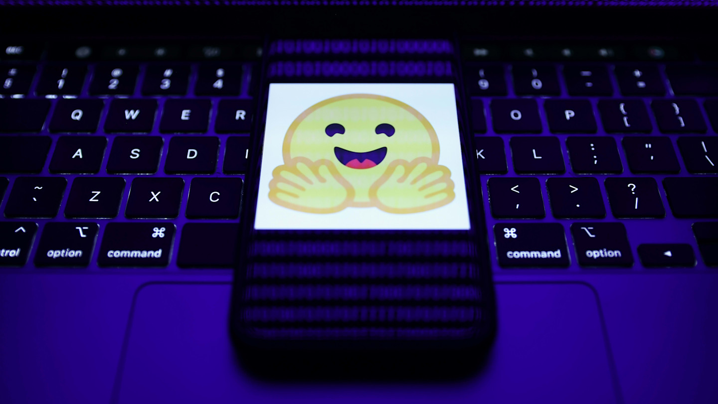 Hugging Face logo displayed on a phone screen over a laptop keyboard, with binary code visible on the laptop screen.