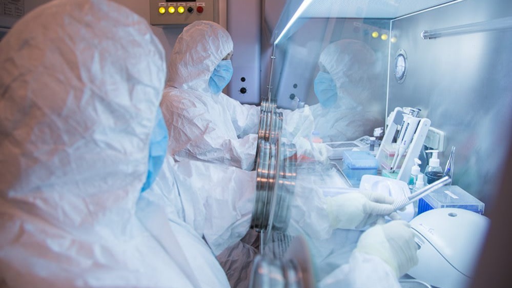 Institute workers in full protective suits and masks work with laboratory equipment behind glass barriers.