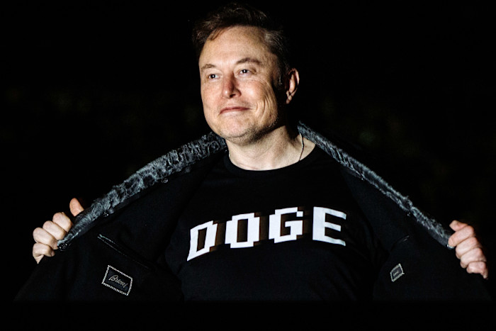 Elon Musk opens his coat to reveal a shirt with the word "DOGE" printed in bold block letters.