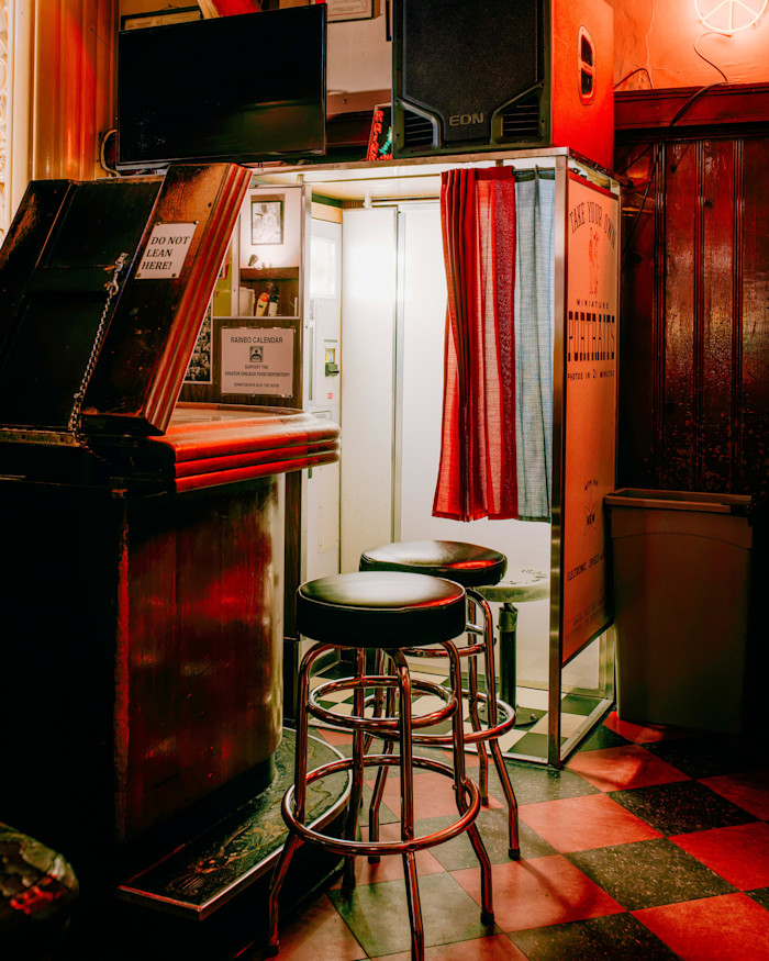 A retro-style photo booth with a red and blue curtain is set up in a dimly lit bar, with two empty stools in front.