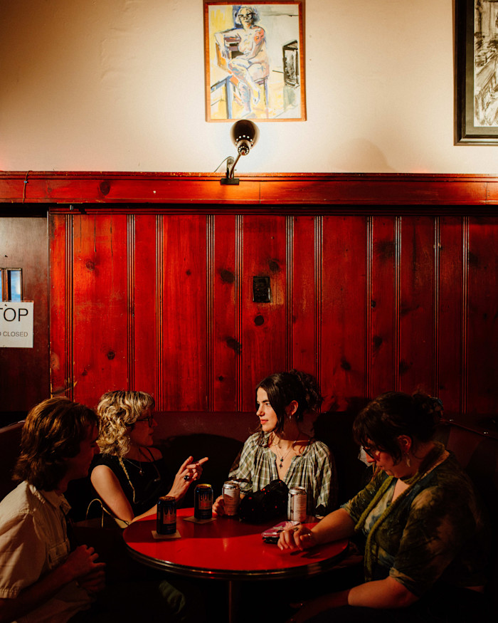 Four people sit around a red table with drinks at Rainbow Club bar, talking in a warmly lit booth beneath wood-paneled walls and a painting.