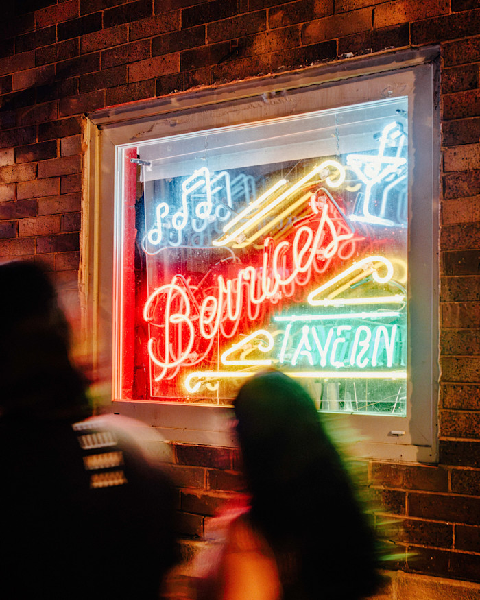 Neon sign reading “Bernice’s Tavern” in a window, glowing at night with blurred figures passing in the foreground.
