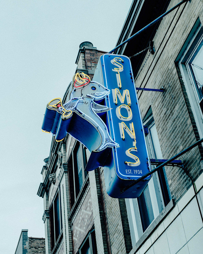 Neon sign for Simon’s Tavern shaped like a fish holding a martini glass, mounted on the exterior brick wall of a building.