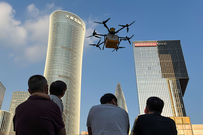 A group of people, including an adult holding a child, watch a Meituan food delivery drone flying between tall buildings in Shenzhen